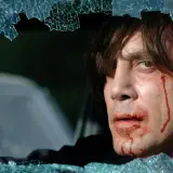 Javier Bardem w filmach, z zakrwawioną twarzą, patrzy przez pękniętą szybę.