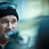 Mężczyzna w czapce i okularach, przypominający Davida Finchera, twórcę mrocznych filmów, patrzy w bok z lekkim uśmiechem.