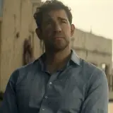 Jack Ryan w niebieskiej koszuli, zamyślony, na tle pustynnego miasta.