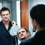 Cezary Pazura w czarnym garniturze, z charakterystyczną bródką, spogląda w lustro. Scena jak z jego filmów.