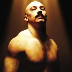 Tom Hardy jako Bronson, w stylu filmów Charlesa Bronsona. Zgolona głowa, wąsy i blizny.