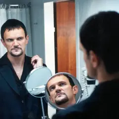 Cezary Pazura w czarnym garniturze, z charakterystyczną bródką, spogląda w lustro. Scena jak z jego filmów.