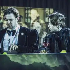 Dwóch mężczyzn przy stole, jeden w czarnym aksamitnym fraku, drugi w mrocznej szacie. Scena z "Ofelii", gdzie napięcie wisi w powietrzu.