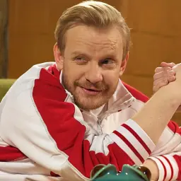 Bartosz Żukowski w sportowej bluzie, z uśmiechem na twarzy, w scenie przypominającej siłowanie na rękę. Znany z wielu filmów, seriali i programów.