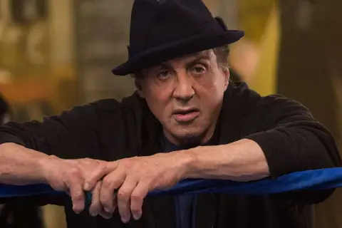 Sylvester Stallone jako Rocky Balboa, w czarnym kapeluszu i bluzie, opiera się o linę ringu.