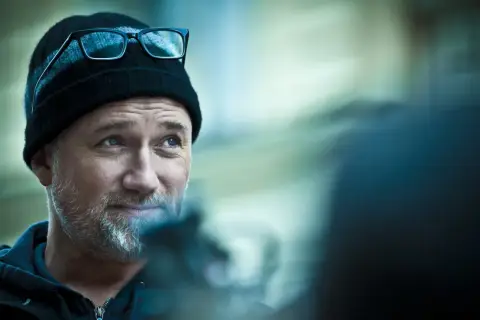 Mężczyzna w czapce i okularach, przypominający Davida Finchera, twórcę mrocznych filmów, patrzy w bok z lekkim uśmiechem.