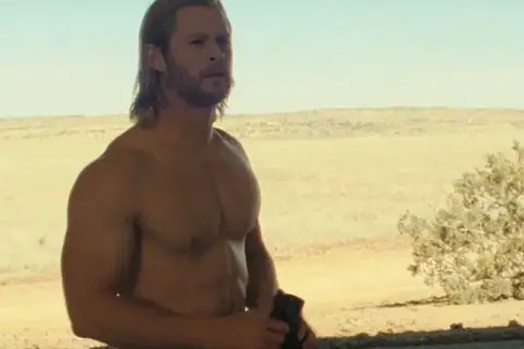 Chris Hemsworth w jednym z filmów, z rozwianymi blond włosami i wspaniałą muskulaturą, stoi na tle pustynnego krajobrazu.