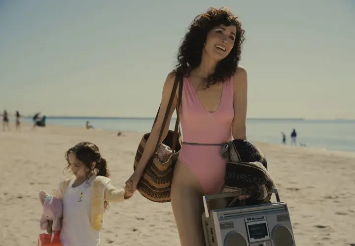 Rose Byrne w różowym kostiumie kąpielowym na plaży z córką, niosąc torbę i boombox.