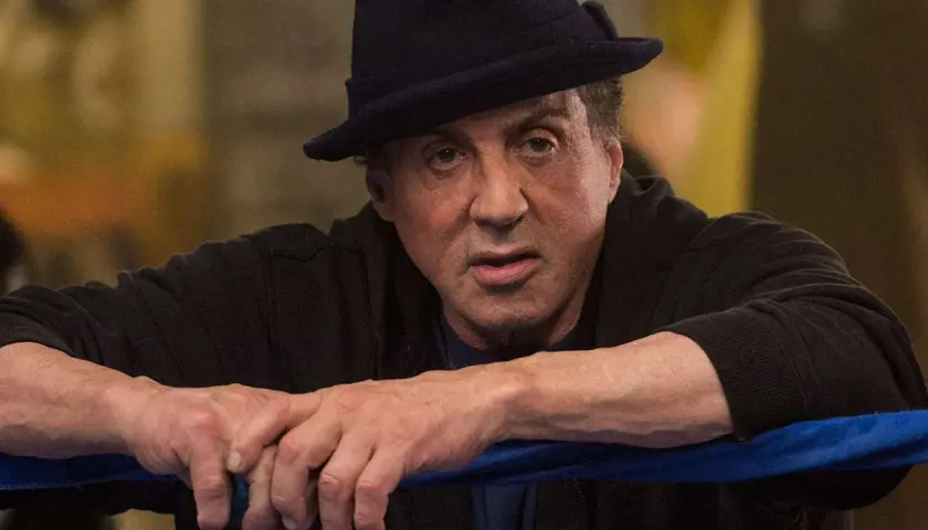 Sylvester Stallone jako Rocky Balboa, w czarnym kapeluszu i bluzie, opiera się o linę ringu.