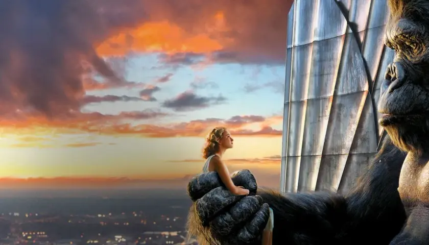 King Kong trzyma kobietę na dłoni, patrząc na zachód słońca nad miastem.