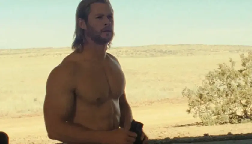 Chris Hemsworth w jednym z filmów, z rozwianymi blond włosami i wspaniałą muskulaturą, stoi na tle pustynnego krajobrazu.