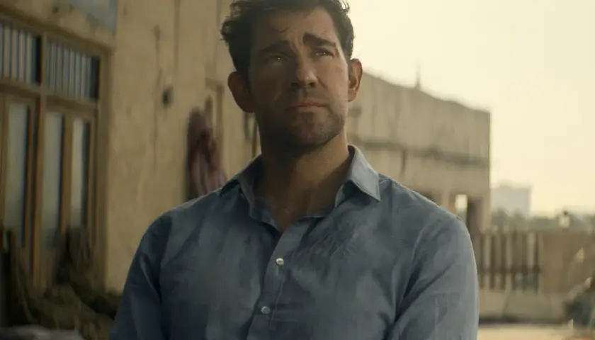 Jack Ryan w niebieskiej koszuli, zamyślony, na tle pustynnego miasta.