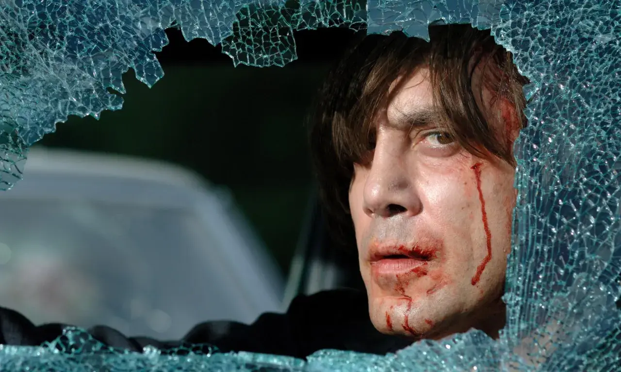 Javier Bardem w filmach, z zakrwawioną twarzą, patrzy przez pękniętą szybę.