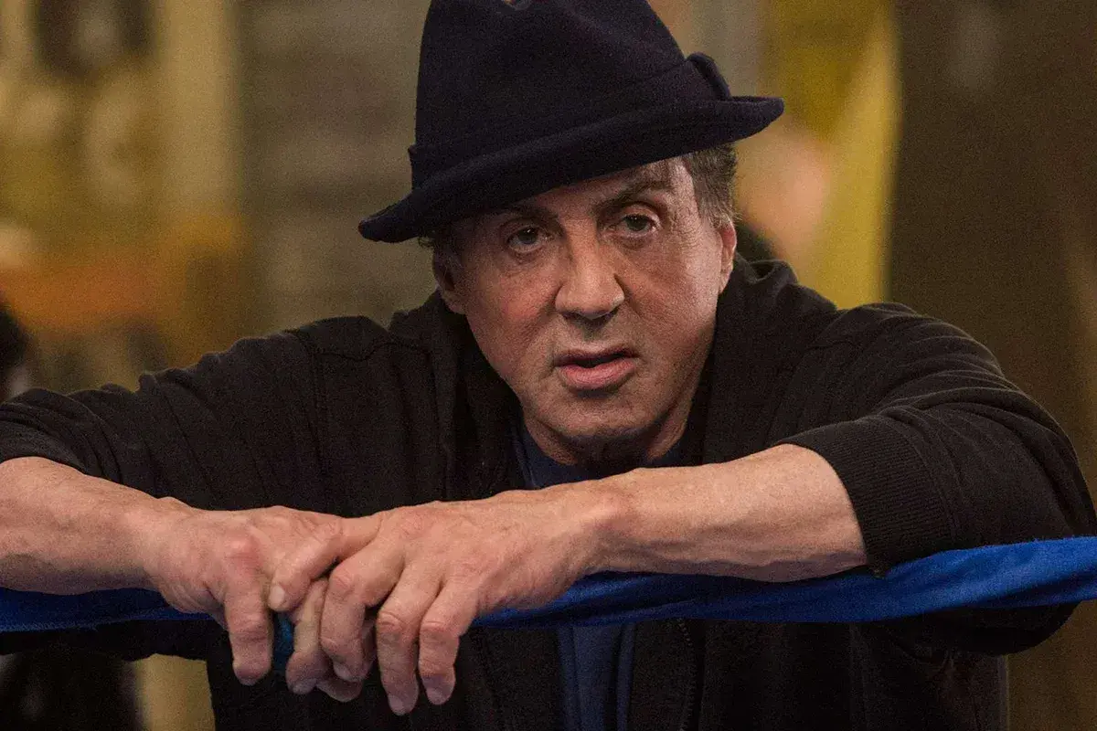 Sylvester Stallone jako Rocky Balboa, w czarnym kapeluszu i bluzie, opiera się o linę ringu.