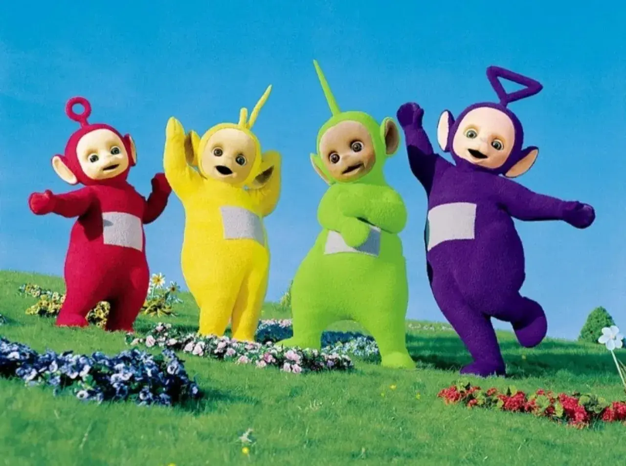 Tinky Winky, Dipsy, Laa-Laa i Po, czyli Teletubisie, machają radośnie na zielonej łące.