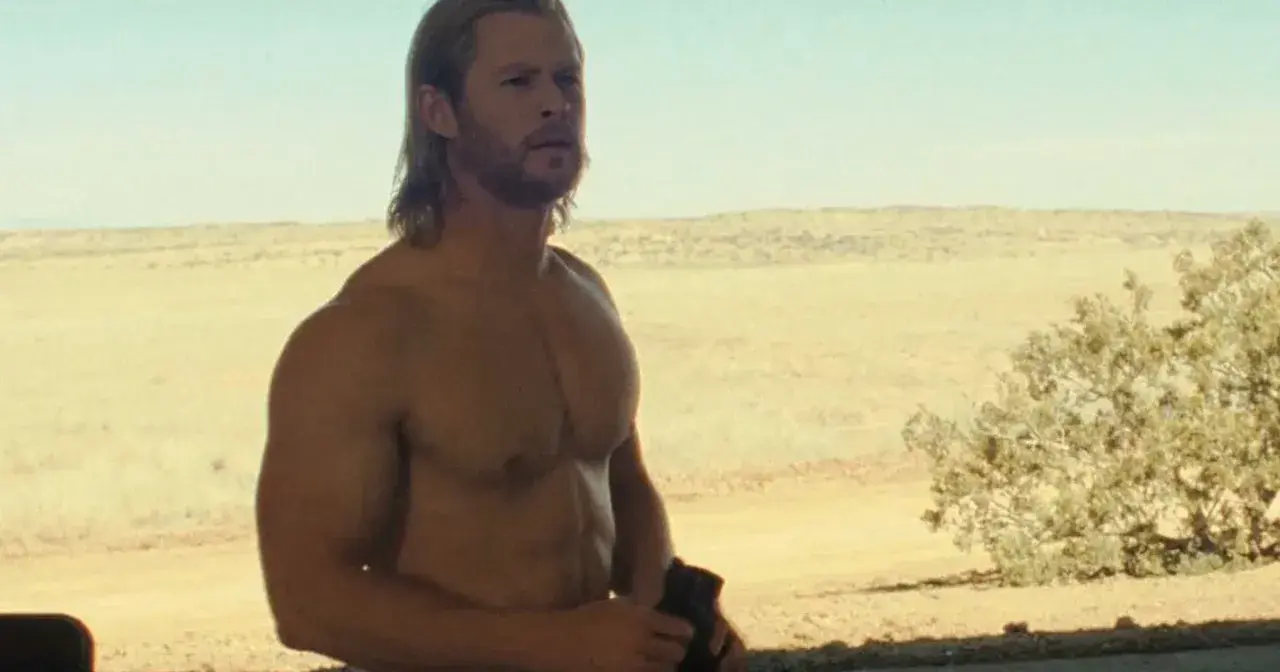 Chris Hemsworth w jednym z filmów, z rozwianymi blond włosami i wspaniałą muskulaturą, stoi na tle pustynnego krajobrazu.