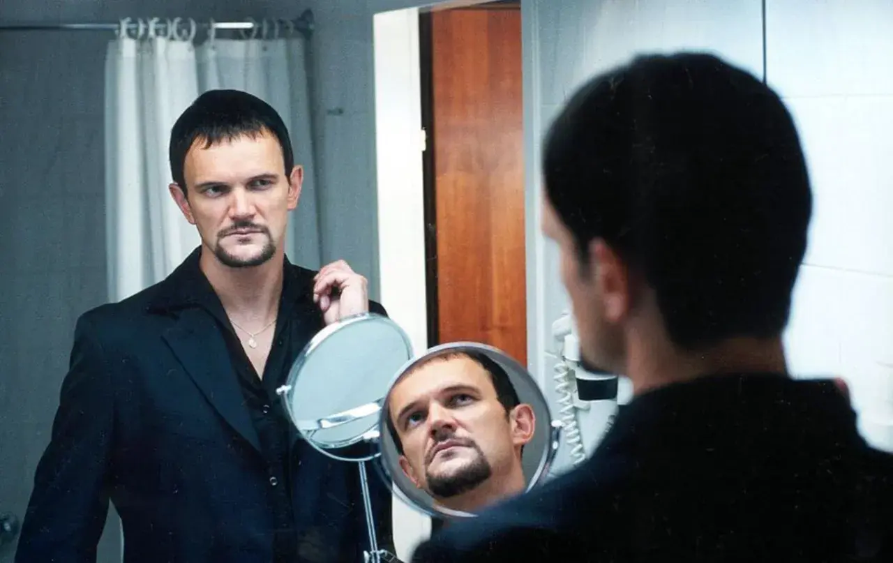 Cezary Pazura w czarnym garniturze, z charakterystyczną bródką, spogląda w lustro. Scena jak z jego filmów.