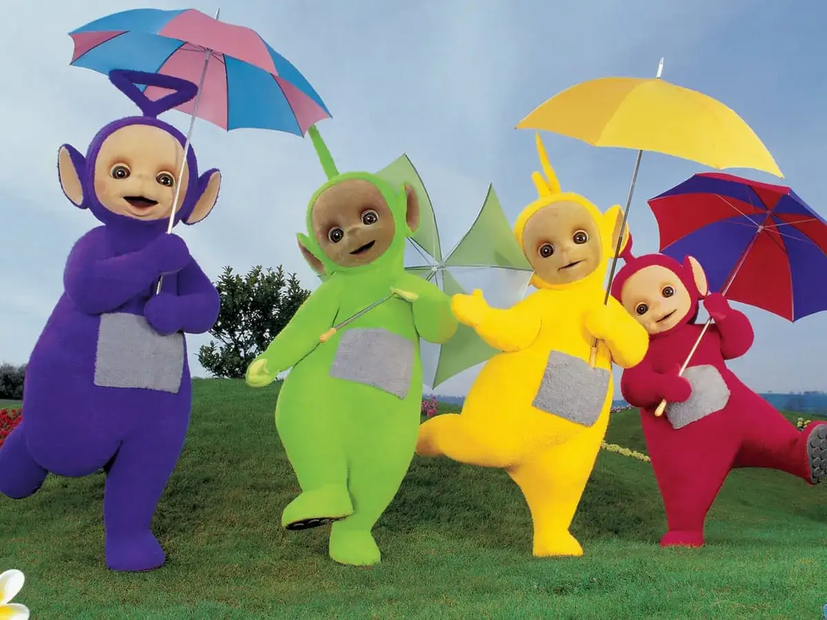 Tinky Winky, Dipsy, Laa-Laa i Po, czyli teletubisie imiona, tańczą z parasolkami na trawie.
