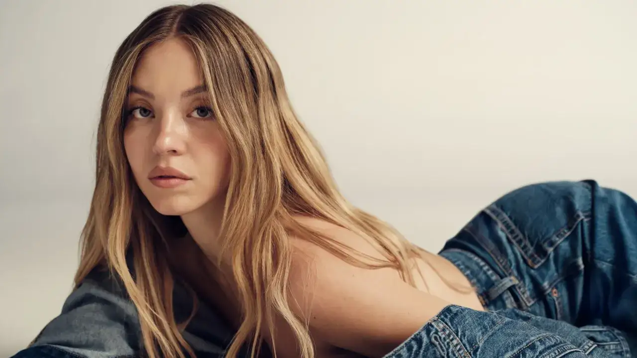 Sydney Sweeney w dżinsowej kurtce, z rozwianymi blond włosami, patrzy wprost na widza.