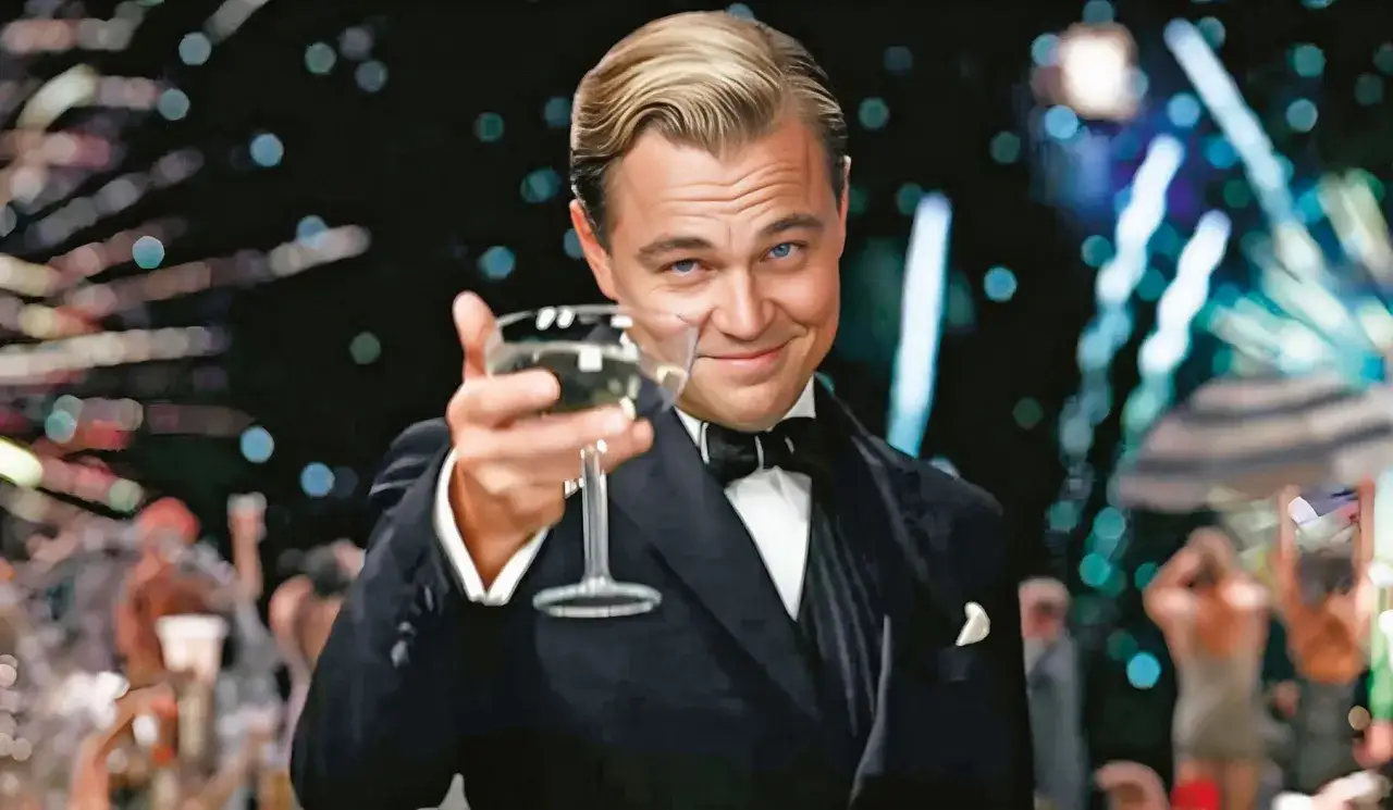 Jay Gatsby wznosi toast kieliszkiem szampana, uśmiechając się do Ciebie na tle rozświetlonej, imprezowej nocy.