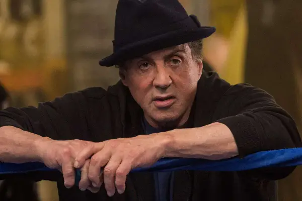 Sylvester Stallone jako Rocky Balboa, w czarnym kapeluszu i bluzie, opiera się o linę ringu.
